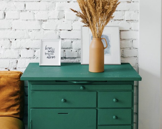 A green drawer against a white brick wall. Pictures in frames and a vase are placed on top of it.